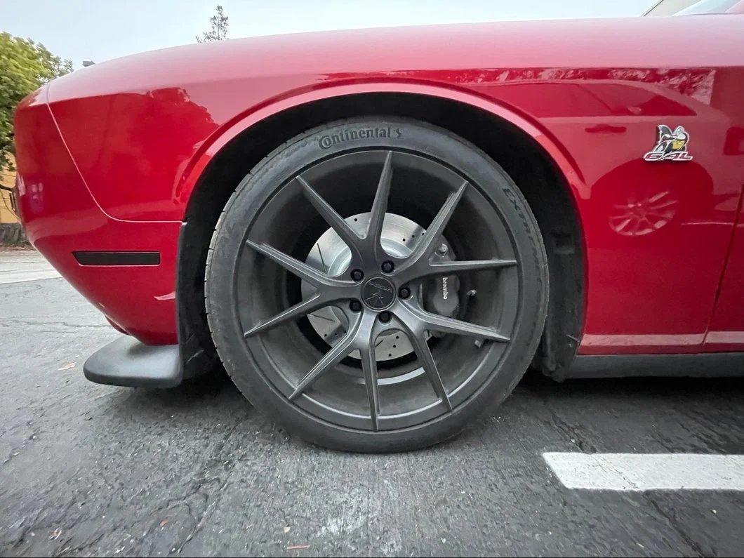 Front wheel of a Dodge Challenger Scat Pack showing a large Brembo brake caliper, drilled brake rotor, Continental tire, and aftermarket performance wheel.