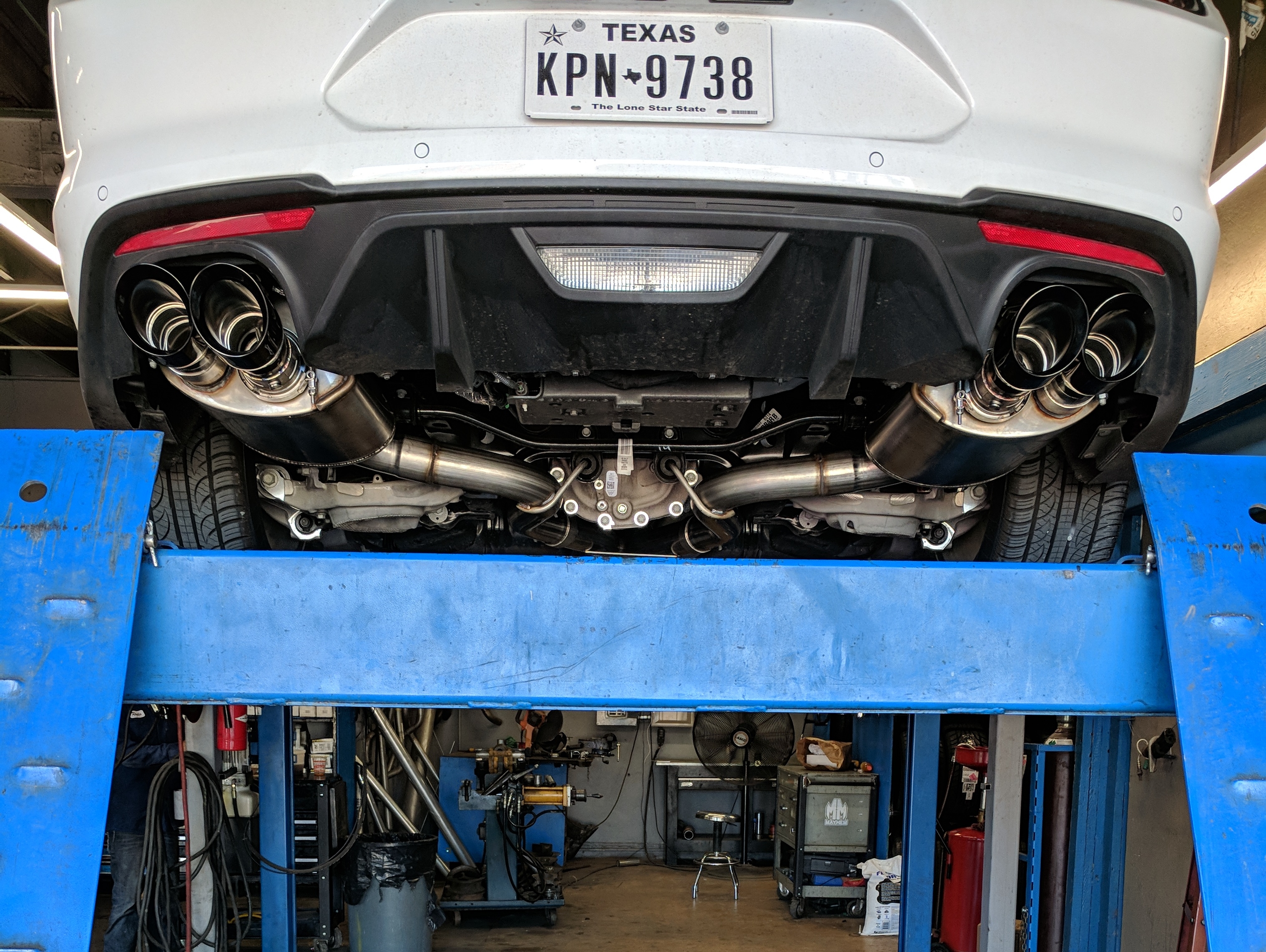 Rear underside view of a late-model Ford Mustang on a vehicle lift, featuring a quad-tip active cat-back exhaust system with polished exhaust tips, performance mufflers, and stainless steel exhaust piping.