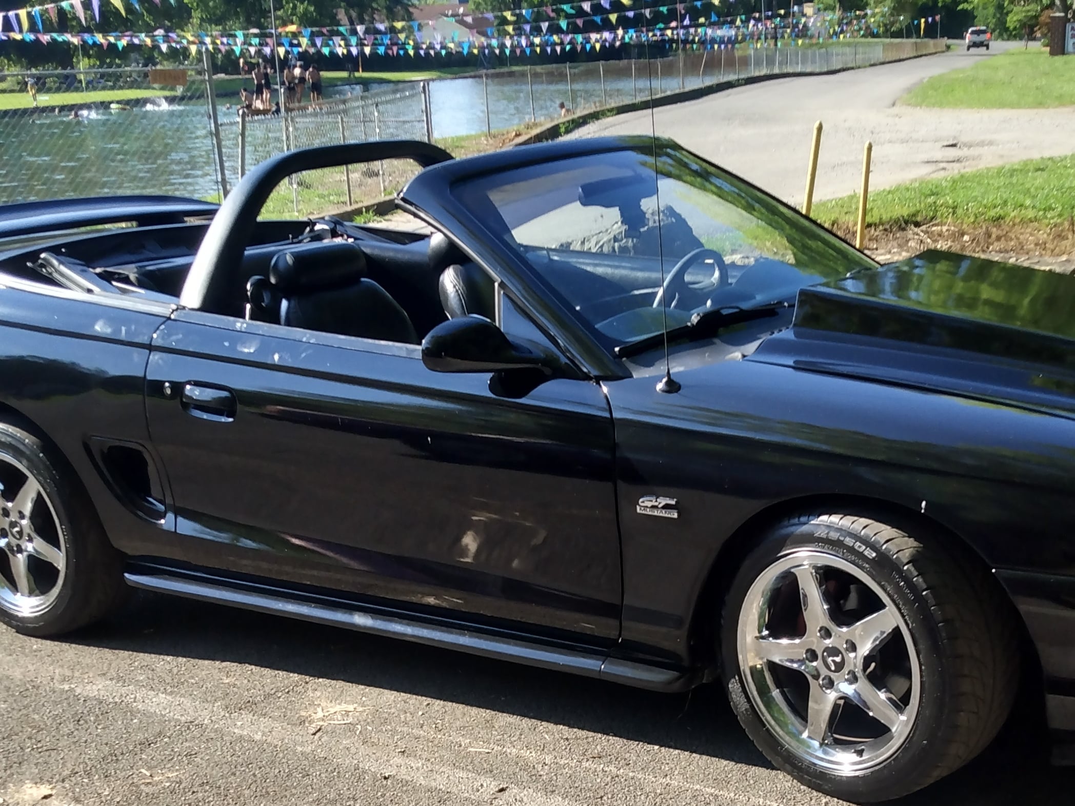 Black SN95 Ford Mustang GT convertible parked near a lakeside recreation area, featuring a raised cowl hood, chrome five-spoke wheels, side scoops, and an aftermarket light bar behind the front seats.