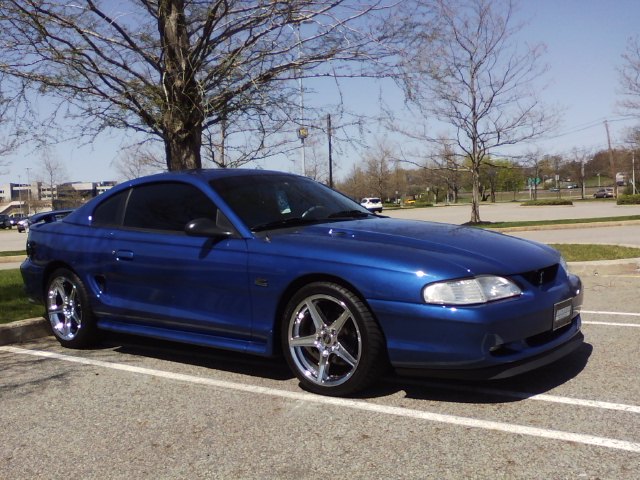 Blue SN95 Ford Mustang GT coupe parked in a parking lot, featuring side scoops, tinted windows, a rear spoiler, lowered suspension, and chrome five-spoke aftermarket wheels.