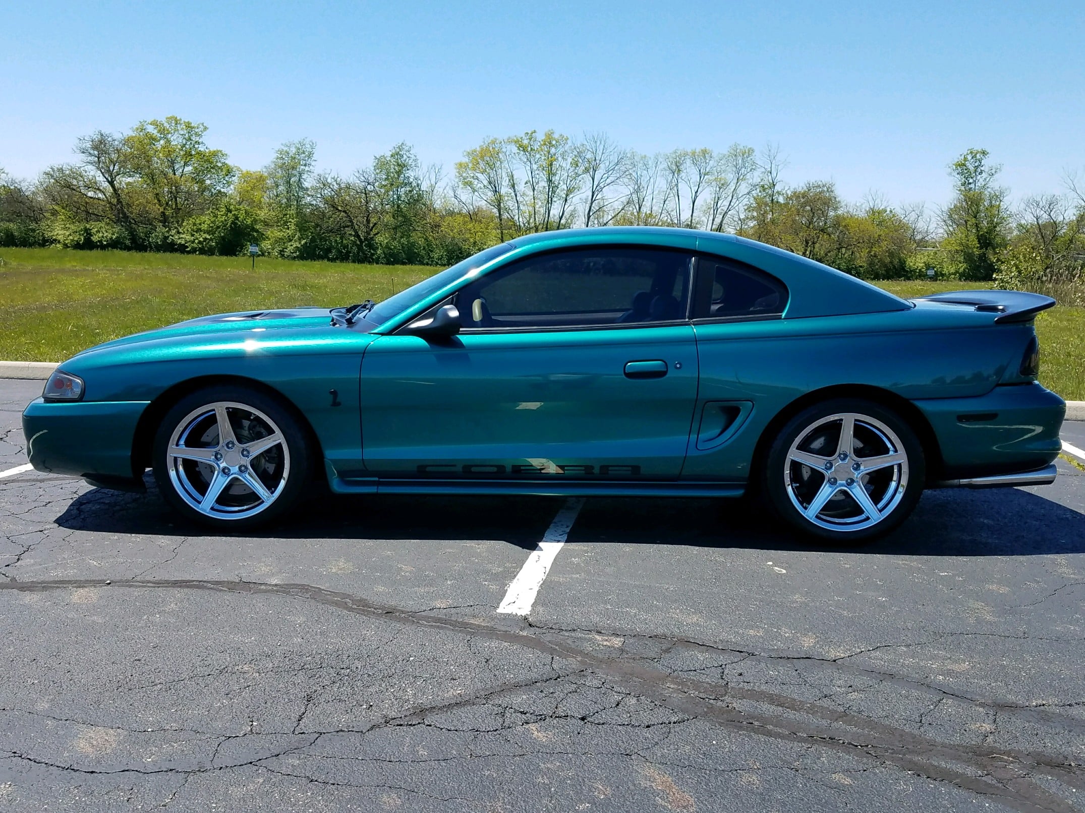 Teal SN95 Ford Mustang Cobra coupe parked in a lot, featuring Cobra fender badging, side scoop bodywork, rear spoiler, tinted windows, and polished five-spoke aftermarket wheels.