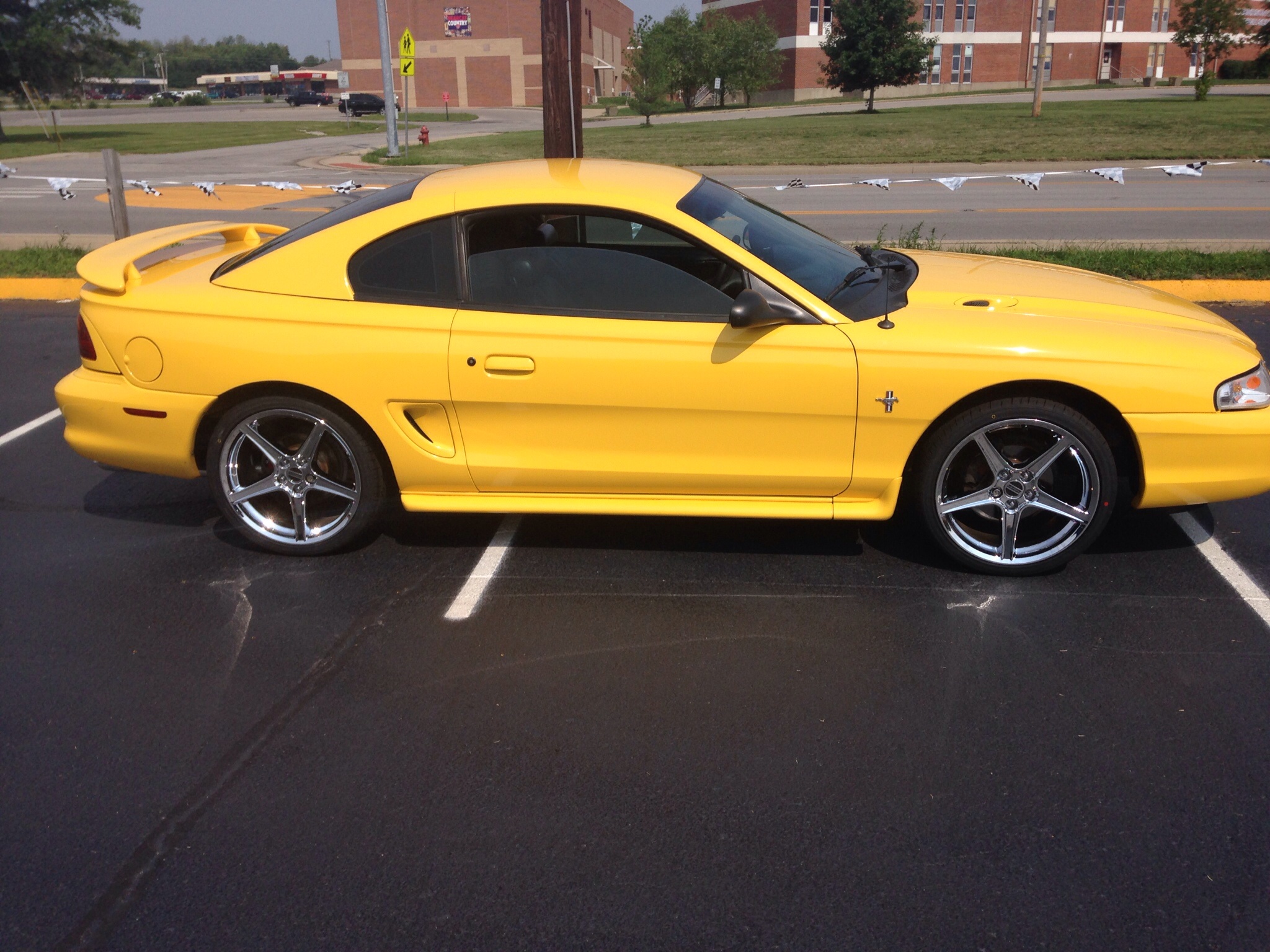 Yellow 1998 SN95 Ford Mustang V6 coupe parked in a lot, featuring side scoops, a rear spoiler, tinted windows, and chrome Saleen Style Chrome Wheels.