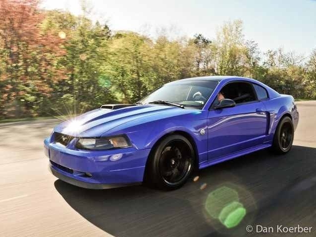 Modified blue 1999&ndash;2004 Ford Mustang New Edge coupe driving on a road, featuring black aftermarket wheels, lowered suspension, and a sporty stance.