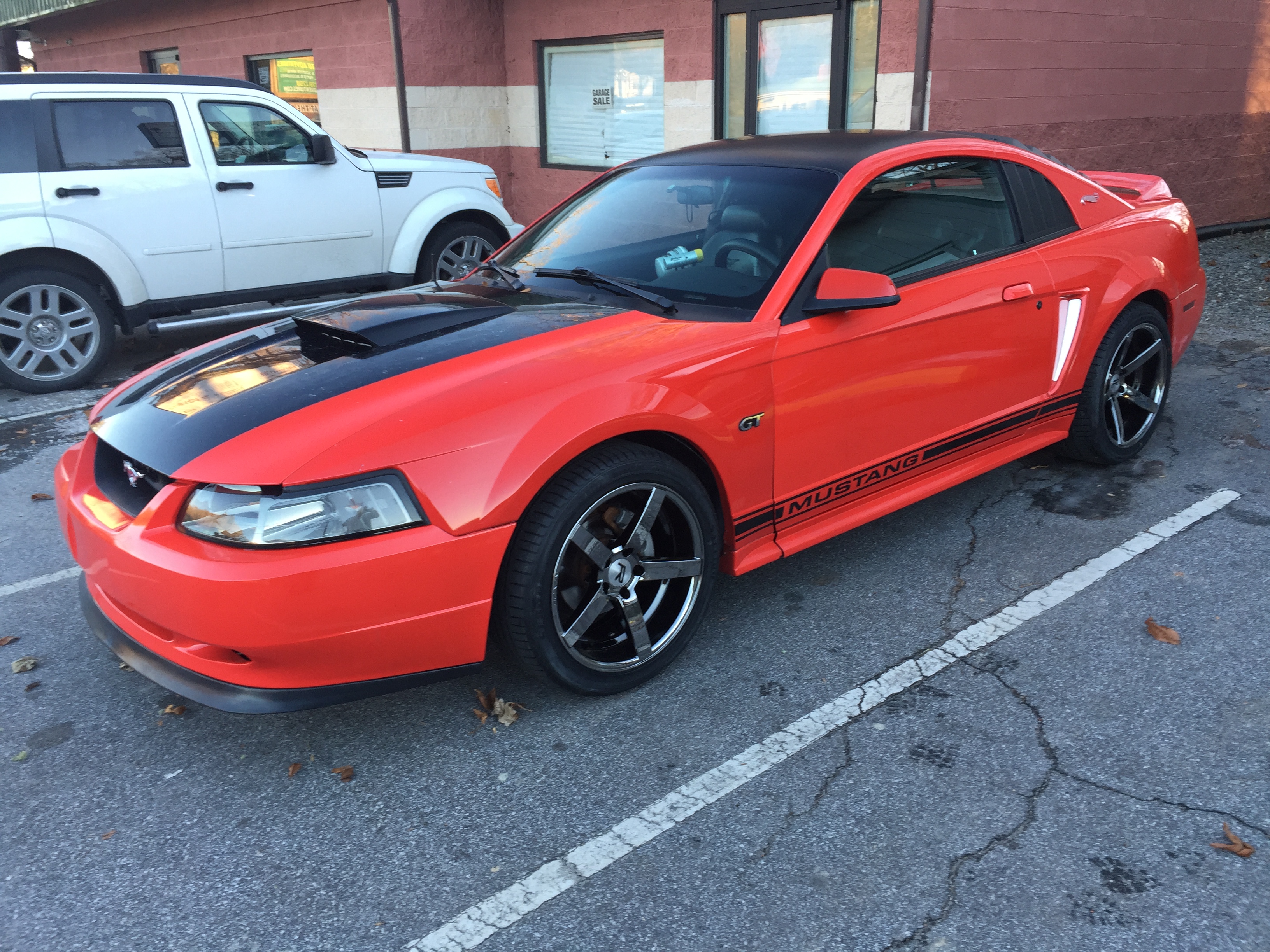 Red 1999&ndash;2004 Ford Mustang GT New Edge coupe with black hood stripe, hood scoop, and black aftermarket wheels parked in a lot.
