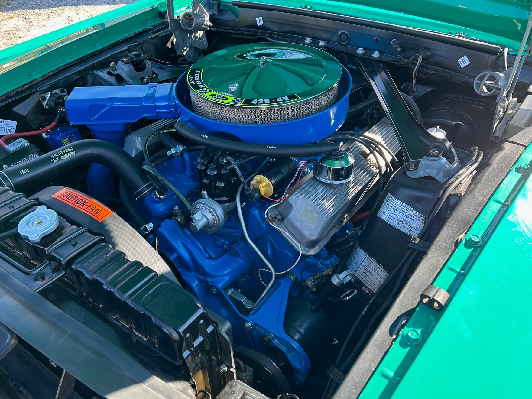 Engine bay of a classic Ford muscle car featuring a blue 428 Cobra Jet V8 engine, chrome air cleaner assembly, finned valve covers, radiator, and factory-style strut tower braces