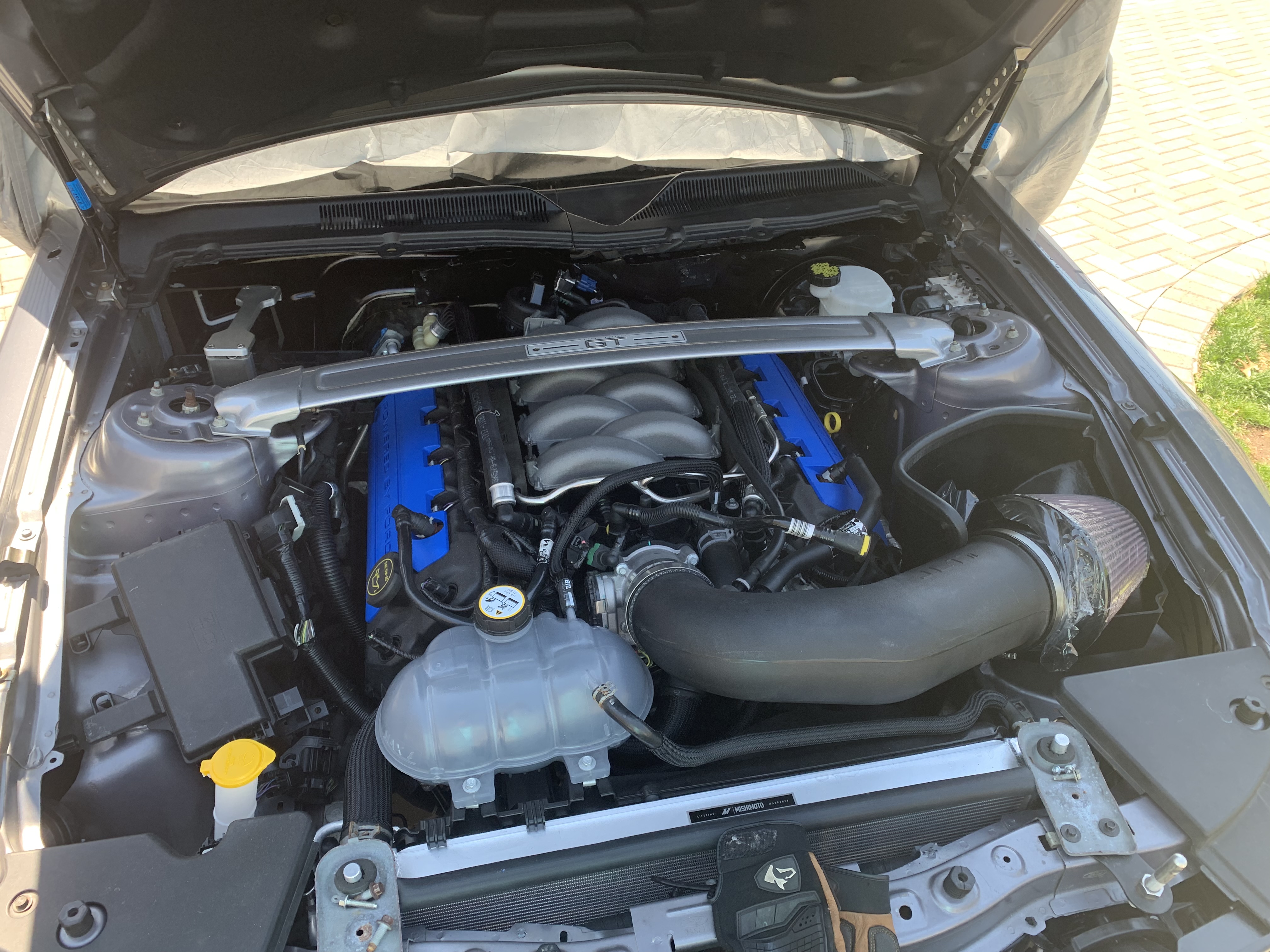 Close-up view of a modern V8 engine bay featuring a cold air intake system, blue valve covers, strut tower brace, upgraded radiator components, and performance cooling hardware installed in a sports car.