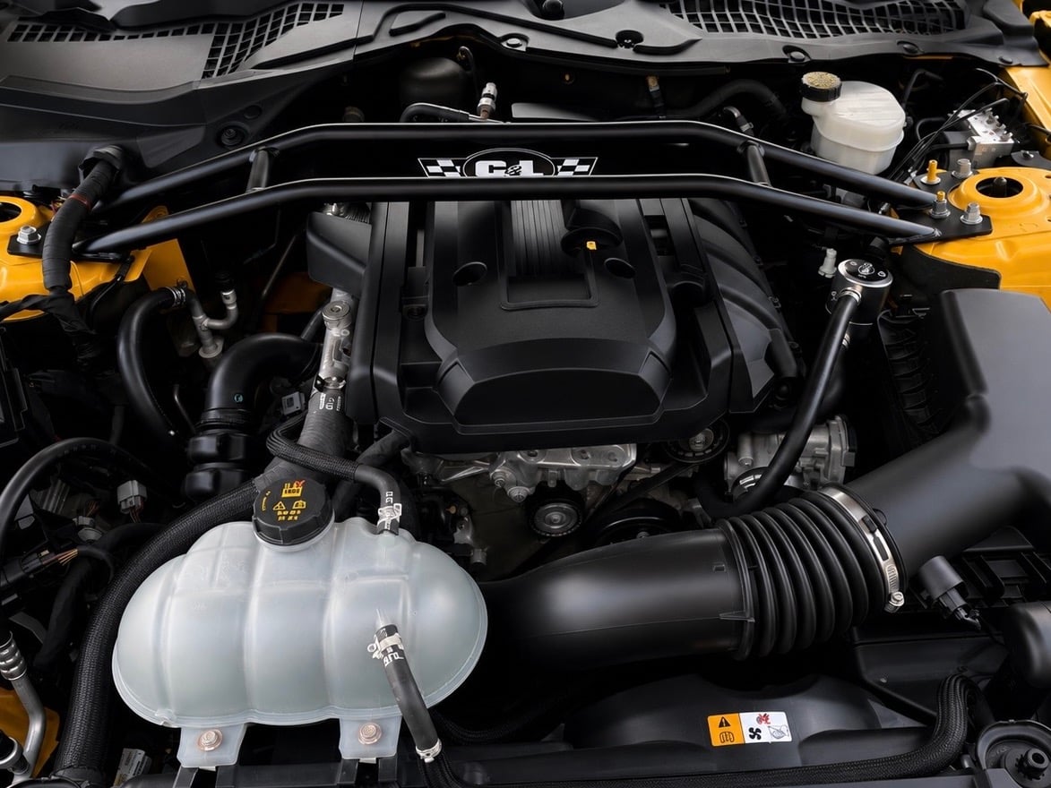 Engine bay of a modern Ford Mustang GT featuring a 5.0L Coyote V8 engine, aftermarket strut tower brace, cold air intake system, coolant reservoir, and factory engine cover.