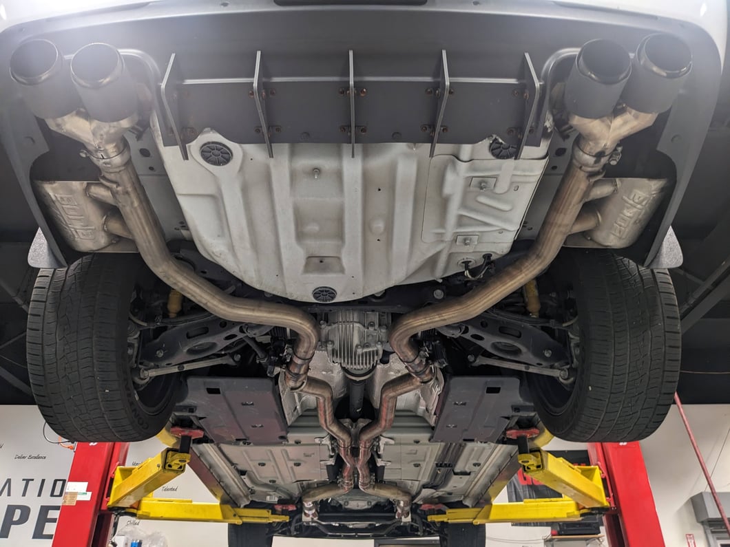 Underside view of a performance car with a dual-exit cat-back exhaust system, featuring stainless steel exhaust piping, rear mufflers, quad black exhaust tips, and an independent rear suspension.